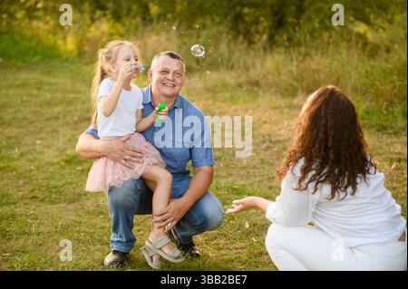 Family having fun in park, talking and playing. Girl happily blowing soap bubbles sitting on her father's lap, mom sitting next to them catching them. Happy childhood concept, summer vacation Stock Photo