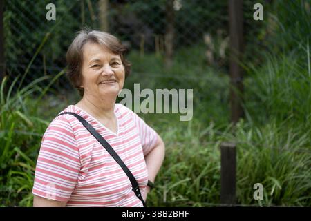 An elderly gray-haired woman in a striped T-shirt stands smiling against the background of a green fence in a summer park. The concept of traveling an Stock Photo