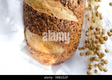 Bread loaf sprinkled with flax and sesame seeds on a white linen tablecloth. Flax seed pods. Sourdough bread. Soft sunlight, evening golden hour. Stock Photo