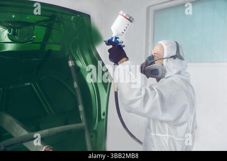 Worker in protective mask and jumpsuit overall spraying base coat of paint on car body elements in painted chamber using professional airbrush. Stock Photo