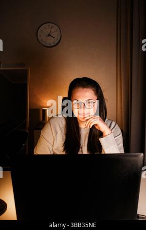 Young woman with eyeglasses working on laptop at home late at night, using modern technologies and getting online education. Remote learning concept. Stock Photo