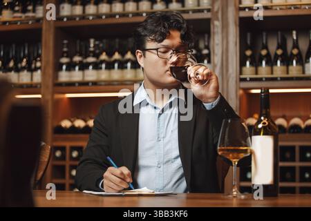 Professional Looking Sommelier Drinking Red Wine in Glass During Tasting Session, Sitting at Table Being in Cellar Stock Photo