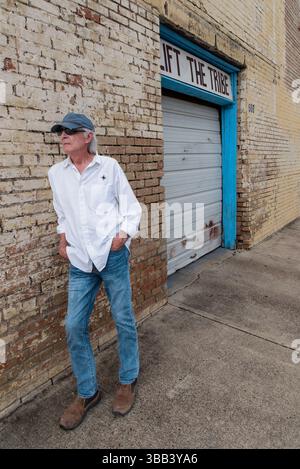 Senior male in blue jeans and white shirt standing leaning against grungy yellow brick wall, words Lift the Tribe in the b.g., McAllen, Texas, USA Stock Photo