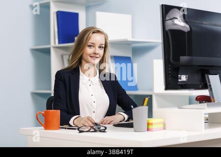 Young pretty business woman sitting at the desk with computer in blue office Stock Photo