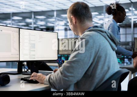 IT engineer looking at PC screen, working in network security data center, ensuring optimal performance. Man supervising server hub, monitoring energy consumption across rigs Stock Photo