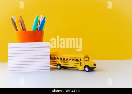 Minimal back to school composition with yellow school bus colorful pens in orange cup and blank lined paper on vibrant yellow background symbolizing Stock Photo