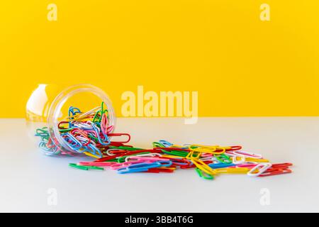 Vibrant paperclips spilling from transparent jar on white and yellow background symbolize creative school supplies visual balance minimal office Stock Photo