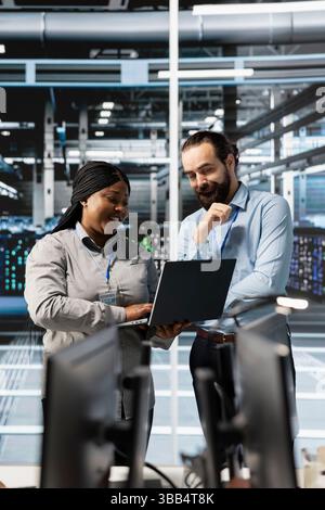 Cheerful team of engineers using laptop in server farm, analyzing data. Jolly teamworking employees examining infrastructure in data center using notebook, ensuring system integrity and security Stock Photo