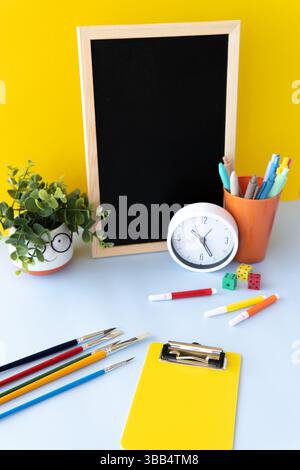 Creative school workspace with blackboard colorful pens markers white clock green plant yellow clipboard and paintbrushes on soft background concept Stock Photo