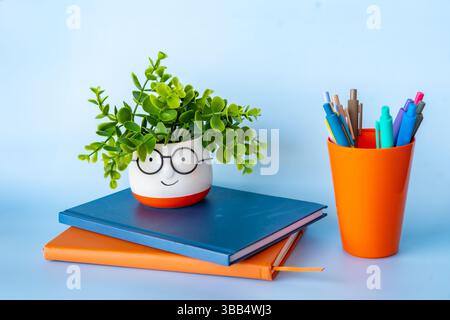 Creative school setup with a smiling plant pot on colorful notebooks and vibrant pens in an orange holder showcasing cheerful learning workspace with Stock Photo