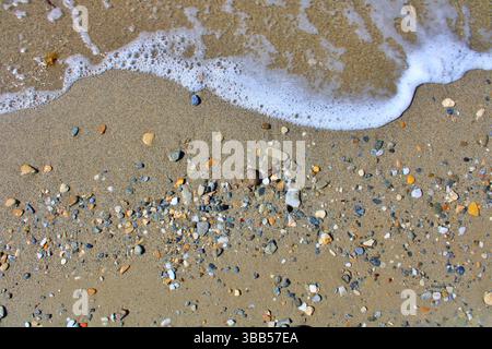 Colorful small stones on sandy beach with sea foam from waves, captured from above on a sunny summer day. Stock Photo