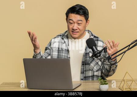 Happy excited blogger Asian man at table recording podcast giving workshop, webinar. Chinese guy influencer speaking into microphone and laptop webcam, making video call discussing on beige background Stock Photo