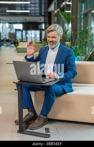 Middle-aged businessman working on laptop computer in modern office lobby talking on online communication video call with employee, boss. Manager man support services looking at webcam of computer. Stock Photo