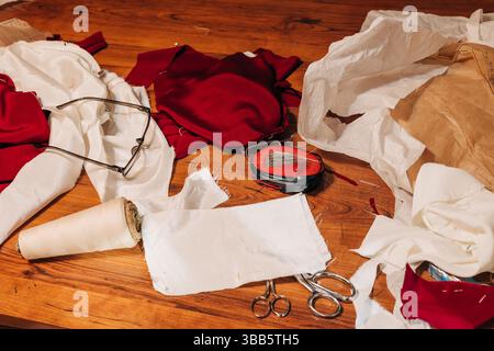 Fabric pieces, scissors, and tools clutter a wooden table in a busy sewing workspace filled with creativity. Stock Photo
