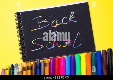 Top view of handwritten on scratch book 'Back to school', colorful crayons and felt-tip pens lined up ascending on yellow background Stock Photo