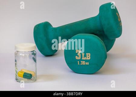 Two blue rubber coated dumbbells are next to glass jar with yellow and green pills on white backgroud. One dumbbell is on top of another dumbbell Stock Photo