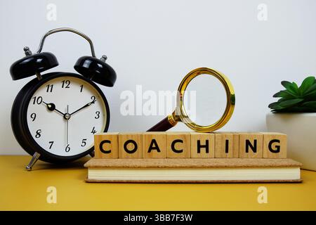 Magnifying glass and alarm clock with wooden blocks alphabet letters COACHING text message on yellow background Stock Photo