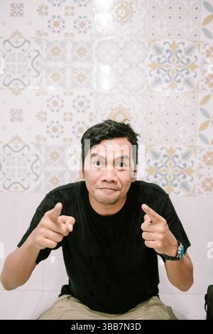 Energetic man with playful gesture, pointing fingers towards the camera with expressive face on patterned tiled wall background Stock Photo