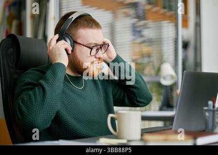 Focused man wearing headphones and eyeglasses, sitting at desk with laptop and cup, adjusting headphones while concentrating on his work indoors with Stock Photo