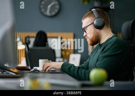 Man with glasses and beard using laptop and wearing headphones in a modern office setting, focused on work with a coffee mug and green object in foreg Stock Photo