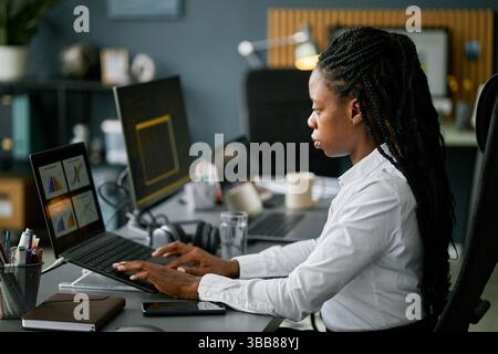 African American woman diligently working at her desk, analyzing data on computer screen while sitting in a modern office setting. She appears focused Stock Photo
