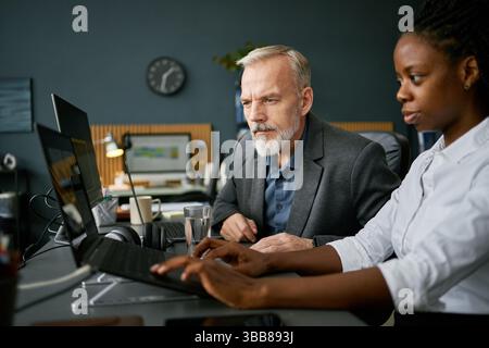 Senior male executive collaborates with young female colleague at modern office, both working on laptops, developing business strategies, and analyzin Stock Photo