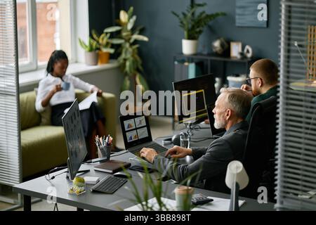 Three colleagues engaged in meeting while discussing project ideas. Office interior with contemporary decor, computer screens, and potted plants provi Stock Photo