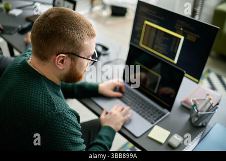 Coder working on project with two monitors and a laptop, immersed in code. Office environment showing programmer focused on software development Stock Photo