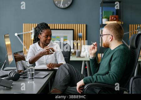 Two colleagues discussing ideas in modern office space with graphs and charts in background. African American woman engaging in conversation with Cauc Stock Photo