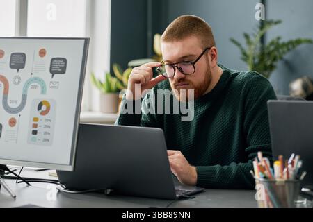 Man with focused expression analyzing data on computer screen in modern office setting with potted plants and mug in background Stock Photo