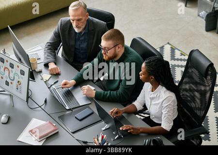 Multiracial team of three, including senior and young professionals, discussing software development project using laptops at modern office desk Stock Photo