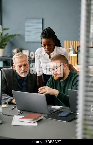 Multiracial business team comprising two men and a woman collaborating on a project in modern office setting, using laptops and other technology devic Stock Photo
