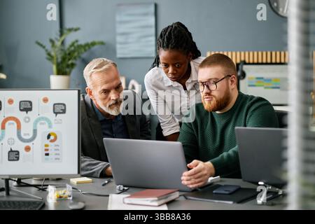 Three colleagues collaborating on project in modern office setting with charts and laptops Two are sitting, one is standing, and all are focused on ta Stock Photo