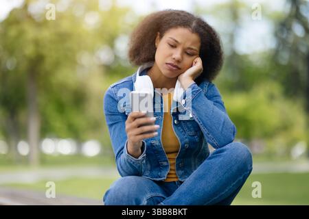 Worried Young Black Female Reading Message On Smartphone While Sitting Outdoors Stock Photo