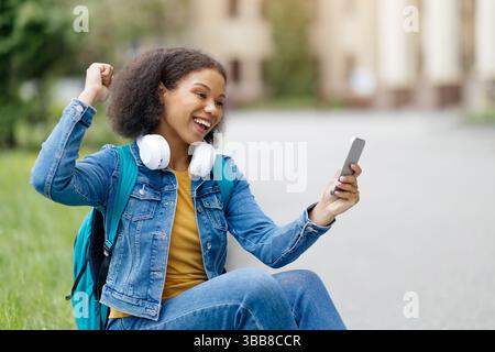 Black female student holding smartphone and celebrating joyfully while sitting on sidewalk Stock Photo