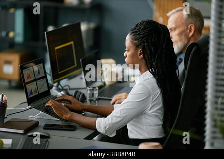 African American woman and Caucasian man working together in professional office environment with laptops and charts on desks, engaging in data analys Stock Photo