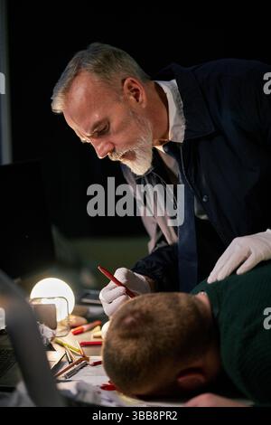 Elderly man concentrating on work by lamplight while younger man sleeping on desk beside him, creating a contrast in work dynamics and energy levels Stock Photo