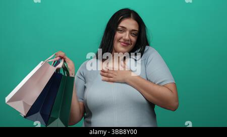 Young woman holding shopping bags smiles confidently against green wall, showcasing plus size fashion and positive shopping experience with a vibrant Stock Photo