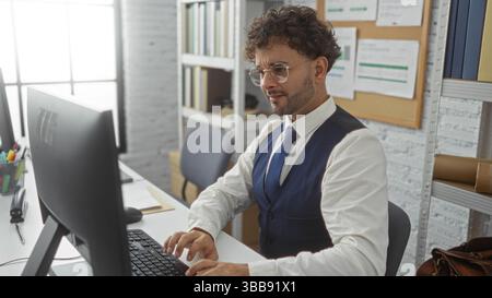 Handsome young hispanic man working indoors at an office desk using a computer, displaying focus and professionalism in a modern workplace environment Stock Photo