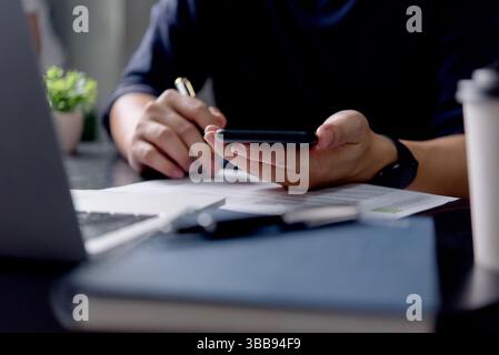 A man is using a cell phone while sitting at a desk. Concept of productivity and focus, as the man is engaged in a task on his phone. The laptop on th Stock Photo