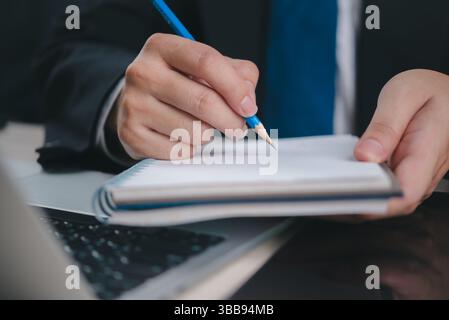 business professional writing in a notebook with a pencil, while working on a laptop. The image symbolizes productivity and business planning in the o Stock Photo