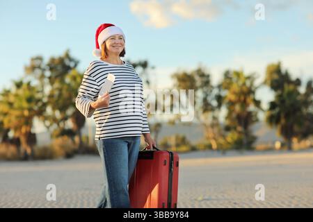 Excited cheerful adult woman in Christmas Santa Claus hat holds red suitcase, passport and boarding ticket walks on beach on seashore with palms, cele Stock Photo