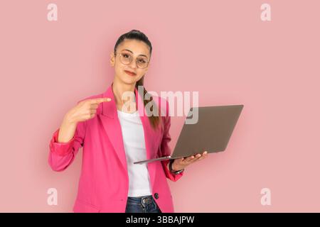business woman holding laptop. Pointing index finger to modern computer pc notebook. Over pink background wearing pink formal jacket, copy space Stock Photo