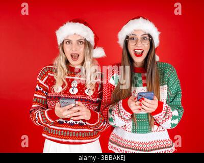 close up portrait of lovely attractive cheerful happy excited Santa ladies. Using mobile cell phone. Wear Santa Claus hat, Christmas sweater. Stock Photo