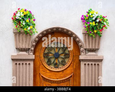 A beautifully crafted wooden door with an ornate circular window, flanked by two decorative stone planters overflowing with vibrant flowers. The backg Stock Photo