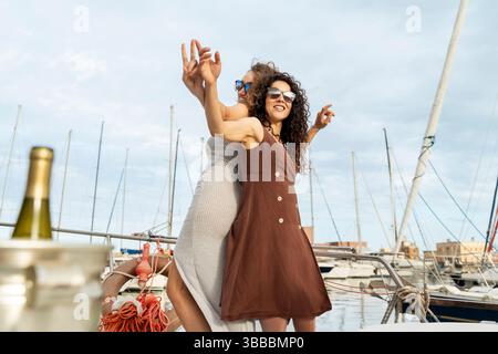 Smiling women in summer dresses dance together on a yacht in the marina, enjoying a carefree holiday party at sunset with boats and masts behind. Stock Photo