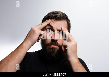 A bearded man points at his forehead with a worried look, suggesting concern, confusion, or perhaps a headache. His expression conveys a sense of unea Stock Photo