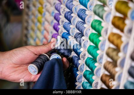 Close-up of hands comparing two spools of dark blue sewing thread to fabric swatch in front of colorful organized thread display Stock Photo