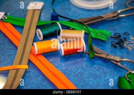 Colorful sewing tools on blue table including green and orange zippers, spools of thread, scissors, and hooks, arranged for garment repair or crafting Stock Photo