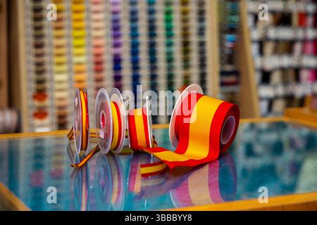 Spain flag - Red and yellow striped ribbon rolls on spools arranged on glass table inside fabric store, with colorful thread displays Stock Photo